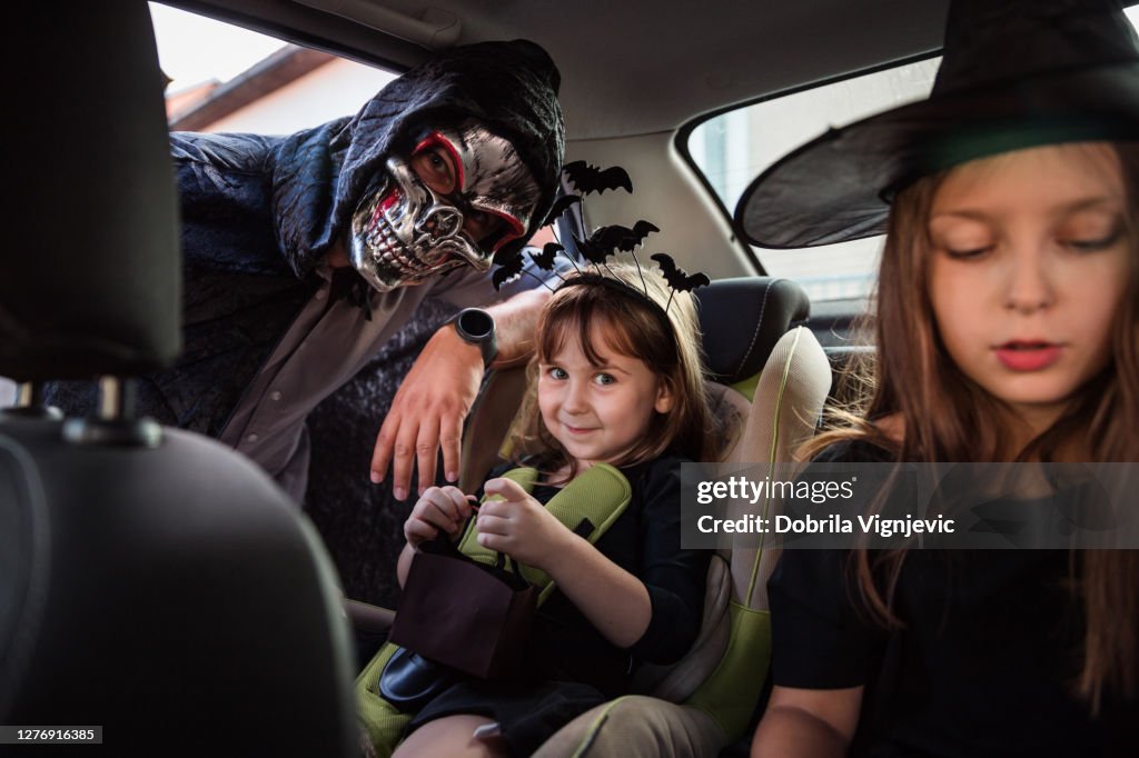 Happy children driving in a car with their dad, all of them wearing Halloween costumes