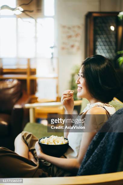 smiling young asian woman enjoying the weekend, watching movie and eating popcorn on the sofa in the living room at home. chilling mood - feet up tv stock pictures, royalty-free photos & images