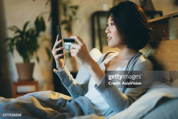 beautiful smiling young asian woman lying on the bed, shopping online on her smartphone and making mobile payment with credit card at home in the evening - all access stock pictures, royalty-free photos & images