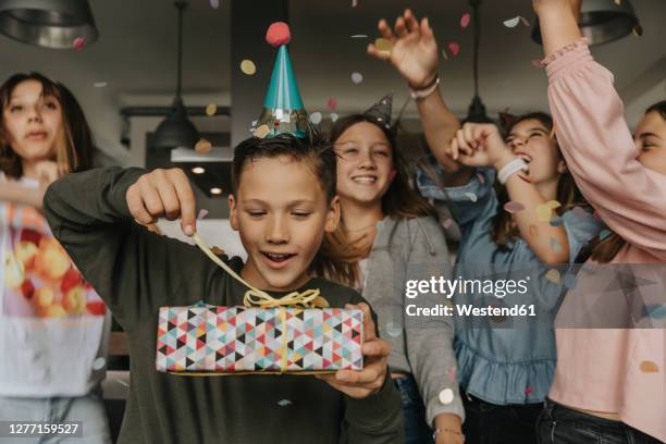 curious birthday boy opening gift while friends dancing in background - verjaardagskado stockfoto's en -beelden