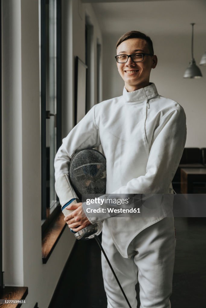 Smiling fencer with foil and mask posing in loft at home