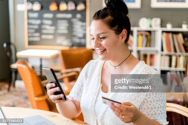 close-up of smiling young woman holding credit card using smart phone in coffee shop - chubby credit fotografías e imágenes de stock