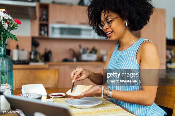 young woman with curly hair buttering bread on table at home - untar de mantequilla fotografías e imágenes de stock