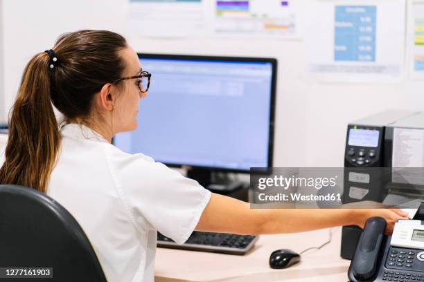 female pharmacist using fax machine on desk while sitting in hospital - faxmachine stockfoto's en -beelden