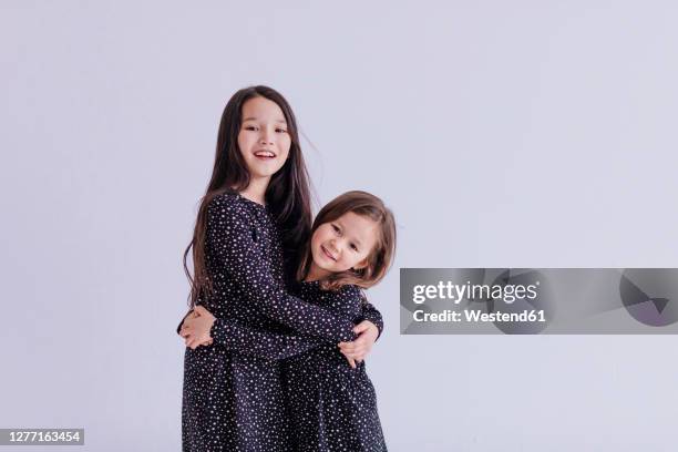 smiling sisters embracing while standing against white background in studio - sesion fotografica fotografías e imágenes de stock