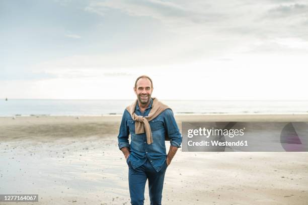 cheerful mature man with hands in pockets walking against sea during sunset - man front view stock pictures, royalty-free photos & images