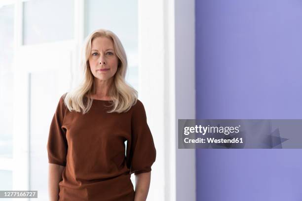 confident female entrepreneur standing against window in office - portrait woman 50 serious stockfoto's en -beelden