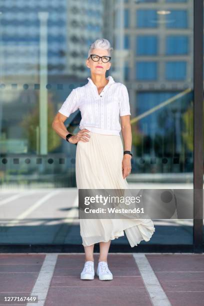 senior woman standing with hand on hip against glass window in city - windy skirt - fotografias e filmes do acervo