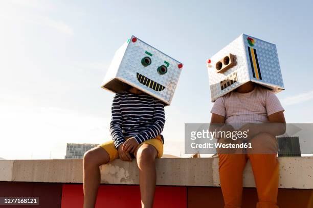friends wearing robot masks sitting on retaining wall against clear sky during sunny day - robot costume stock-fotos und bilder