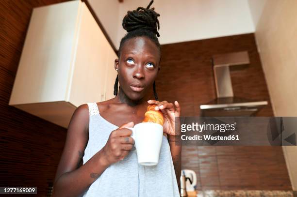 woman looking away while having croissant with coffee in kitchen - dippen stock-fotos und bilder
