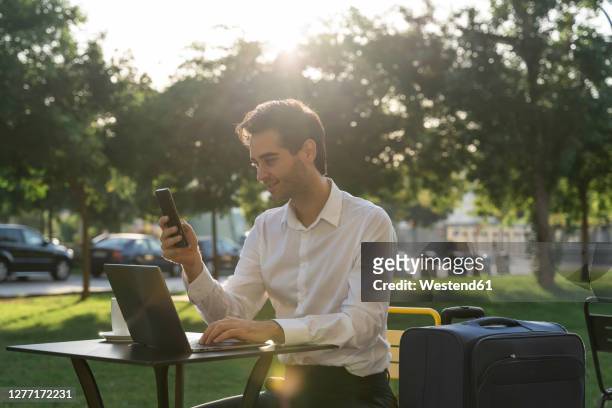 businessman using smart phone and laptop while sitting with suitcase at sidewalk cafe - straßencafe stock-fotos und bilder