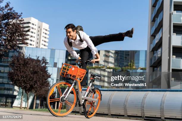 carefree male entrepreneur jumping on electric bicycle against clear sky in city - bicycle stunt stock pictures, royalty-free photos & images