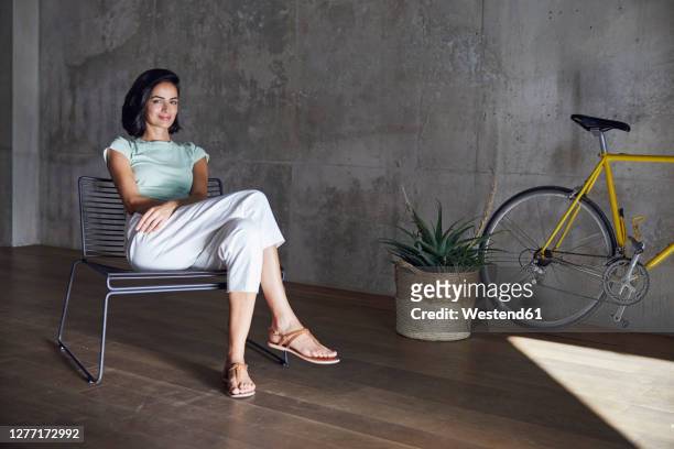 smiling businesswoman relaxing on chair by plant and bicycle in office - sitting chair office relax stock pictures, royalty-free photos & images