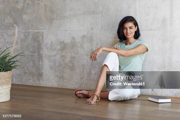 smiling female entrepreneur with book sitting on hardwood floor against wall in office - arab businesswoman with books fotografías e imágenes de stock