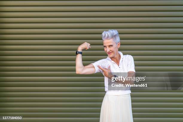 active senior woman pointing at bicep while flexing muscles while standing against shutter - biceps stock pictures, royalty-free photos & images