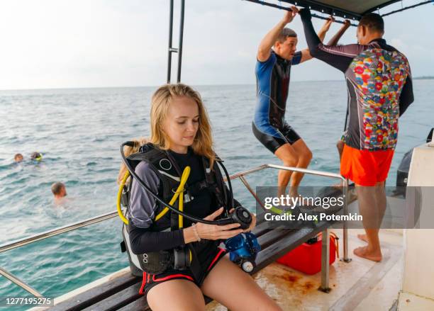 de jonge vrouw die een wetsuit en aqualung draagt wacht op het duiken wanneer de mens voorbereidingen treft om in het overzees van een raad van een boot in de achtergrond te springen. - aqualung diving equipment stockfoto's en -beelden