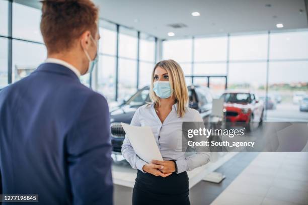 young female car dealer and young male customer with protective face masks talking in a car showroom - car dealership covid stock pictures, royalty-free photos & images