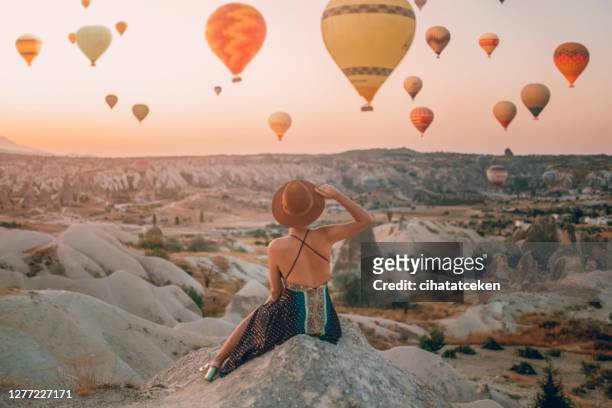 rückblick junge erwachsene frau sitzt auf dem boden und beobachtet luftballons, die auf das tal schauen. kappadokien-sonnenaufgang - cappadocia hot air balloon stock-fotos und bilder