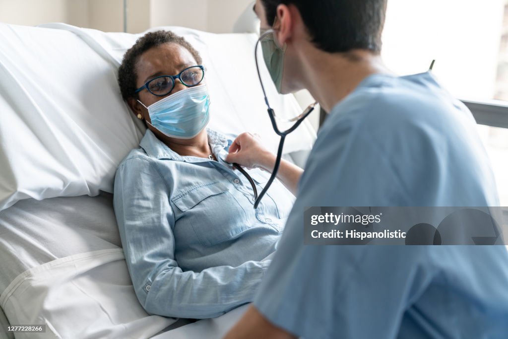 Latin american male nurse checking black female patient's heart with a stethoscope both wearing protective facemasks