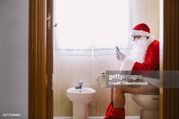 santa claus in the bathroom, sitting on the toilet pooping and looking at his cell phone - bad christmas stock-fotos und bilder