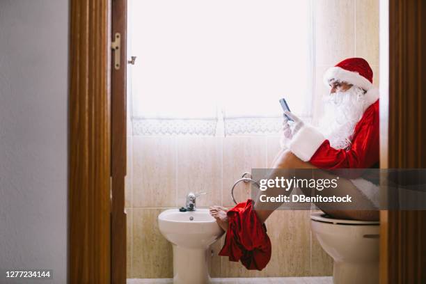 santa claus in the bathroom, sitting on the toilet pooping, his feet propped up on the bidet and looking at his mobile phone. - people peeing stockfoto's en -beelden