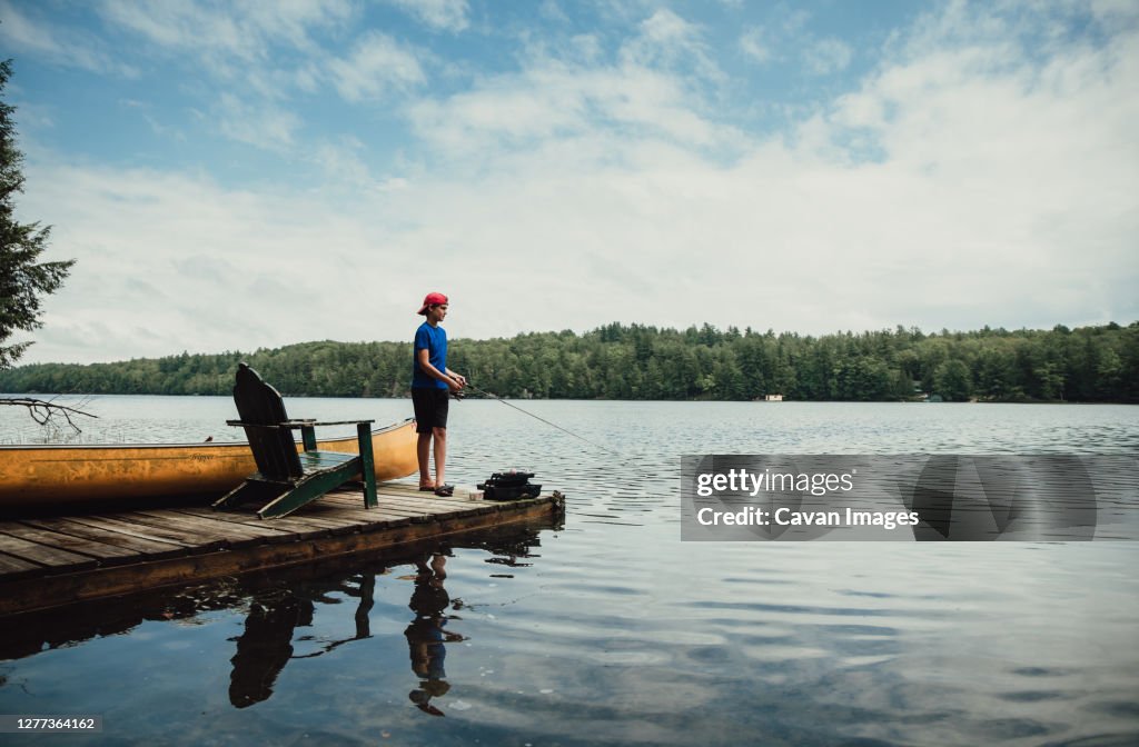 Teen boy fishing from a dock on a calm lake in the summer.
