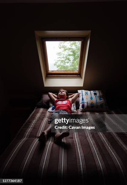 young boy laying on bed looking up through a sky light in a dark room. - skylight stock pictures, royalty-free photos & images