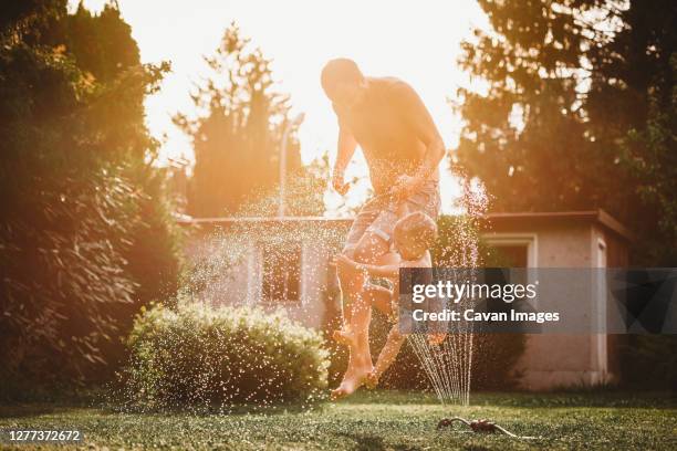 father and child jumping over the water from the sprinkler in garden - jumping sprinkler stock pictures, royalty-free photos & images