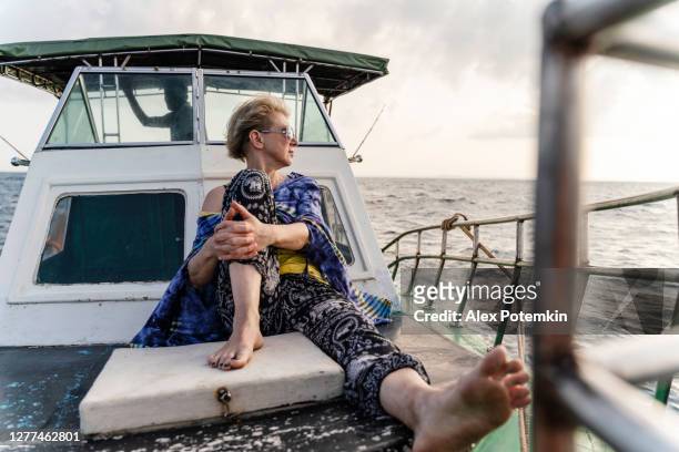 een 50-jarige actieve europese vrouw, een toerist, rust op het dek van een kleine vissersboot tijdens de diepzeevisreis in sri lanka wanneer een kapitein het schip regeert vanuit een dekhuis op de achtergrond. - 50 54 years stockfoto's en -beelden