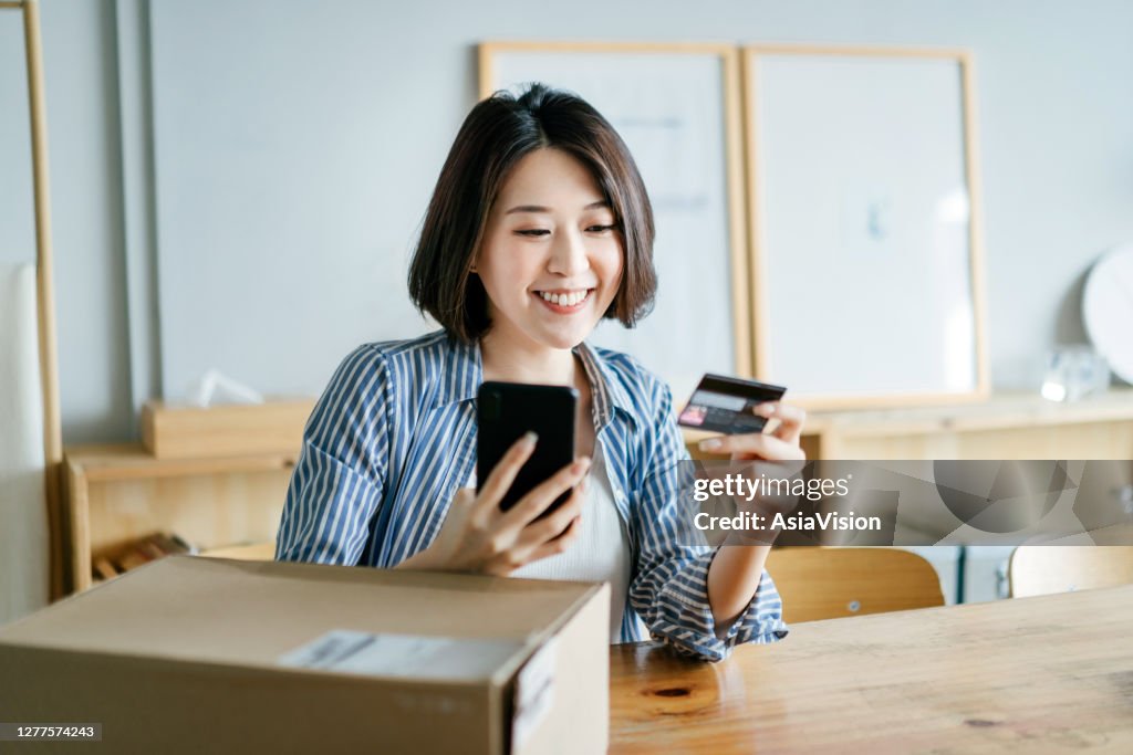 Cheerful young Asian woman shopping online with smartphone and making mobile payment with credit card on hand, receiving a parcel by home delivery service. Technology makes life so much easier