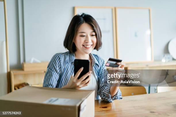 cheerful young asian woman shopping online with smartphone and making mobile payment with credit card on hand, receiving a parcel by home delivery service. technology makes life so much easier - debit cards credit cards accepted stock pictures, royalty-free photos & images