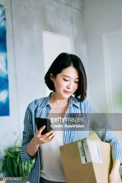 beautiful smiling young asian woman checking smartphone while receiving a parcel at home - returning customer stock pictures, royalty-free photos & images