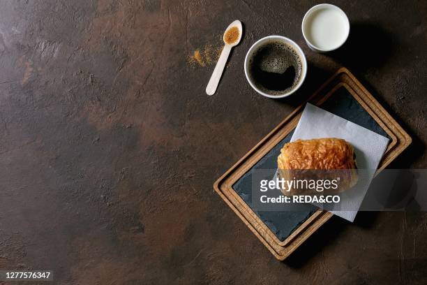 Traditional french puff pastry chocolate bun on slate wooden board with paper cup of coffee and milk. Recycled wooden spoon of cane sugar over dark...