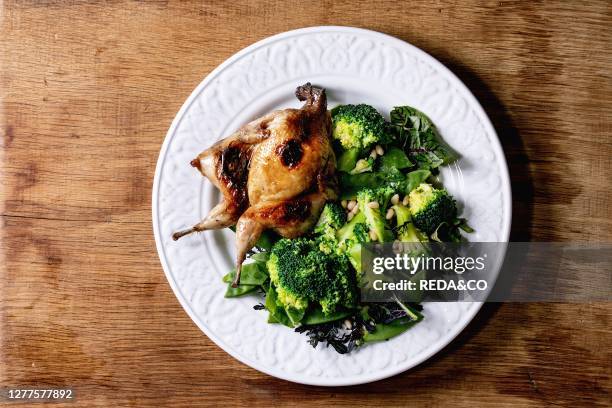 Roasted grilled butterfly quail on ceramic plate with garnish green salad and broccoli over wooden background. Flat lay. Space.
