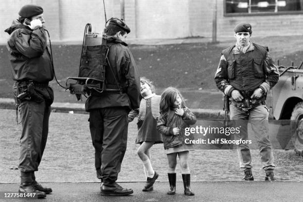 Two young girls stand with members of the British Army's Parachute Regiment, one of whom speaks on a field telephone, in the primarily Catholic,...