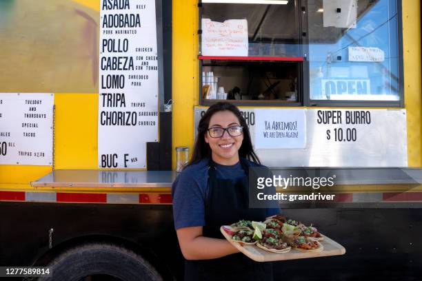 vrij jonge mexicaanse vrouw voor haar kleine bedrijfsvoedselvrachtwagen tacotribune die bij de camera glimlacht en een verscheidenheid van haar heerlijke gastronomische taco's toont - foodtruck stockfoto's en -beelden