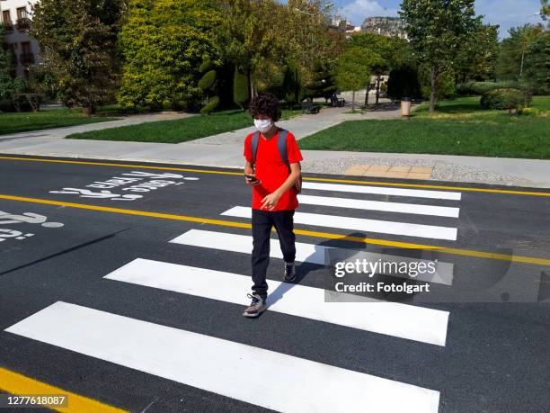 menino de camiseta vermelha atravessando a estrada com máscara facial e celular - travessia de pedestres marca de rua - fotografias e filmes do acervo