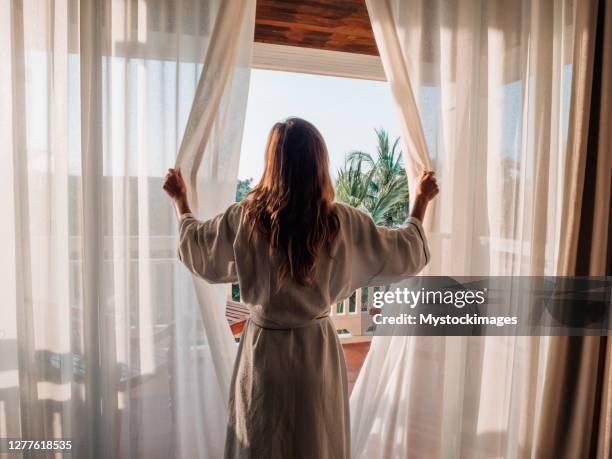 mujer abriendo cortinas en la habitación al amanecer - bathrobe fotografías e imágenes de stock