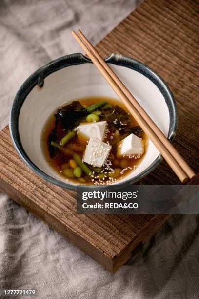Japanese Miso broth soup with silk tofu cubes. Soy beans edamame. Green beans in traditional bowl with chopsticks on wooden japanese table on grey...
