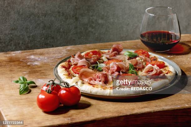 Fresh baking homemade pizza napolitana with prosciutto ham, cheese, tomatoes, basil on plate, glass of red wine over wooden table background. Home...