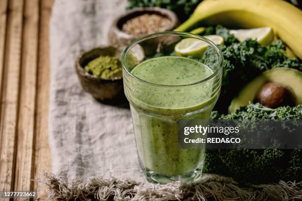 Glass of green healthy vegan smoothie. Glass straw. Ingredients above. Kale. Bananas. Avocado. Lime. Non-diary milk. Matcha powder and seeds over...