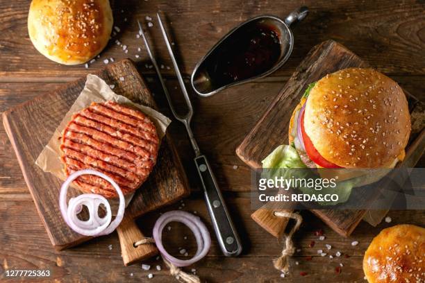 Fresh homemade burger on little cutting board and raw cutlet and sliced onion. Served with ketchup sauce and meat fork over wooden table. Dark rustic...