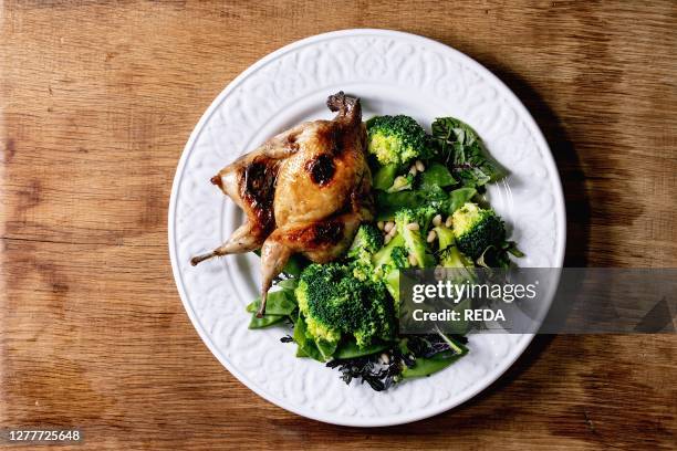 Roasted grilled butterfly quail on ceramic plate with garnish green salad and broccoli over wooden background. Flat lay. Space.