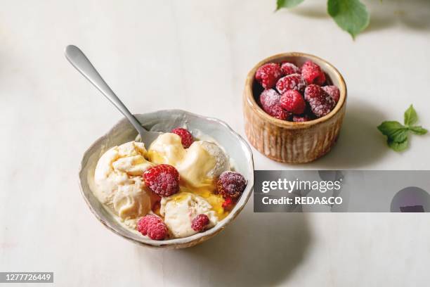 Homemade melted caramel vanilla ice cream with syrup and frozen raspberries in ceramic bowl standing on white marble table.