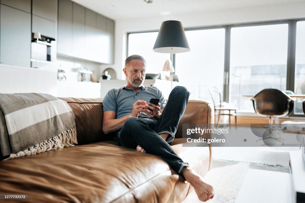 Stattliche bärtige Midaged Mann Blick auf Handy auf sofa