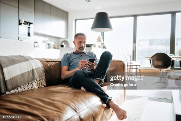 beau homme midané barbu regardant le téléphone portable sur le sofa - barefoot stock photos et images de collection