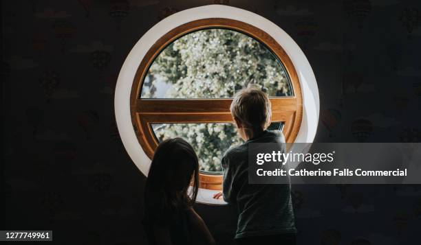 two silhouetted children look out of an unusual circular feature window at sunny tree view. - family with two children british stock pictures, royalty-free photos & images