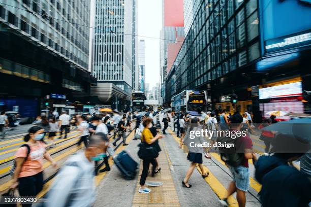 blurred motion of a crowd of busy commuters with protective face mask crossing the street in downtown financial district against contemporary corporate skyscrapers and city traffic during rush hour - crowd masks stock pictures, royalty-free photos & images