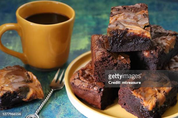 close-up image of homemade chocolate salted caramel brownie squares piled on yellow plate and on slate, metal fork, yellow coffee mug, focus on foreground - salted brownie stock pictures, royalty-free photos & images
