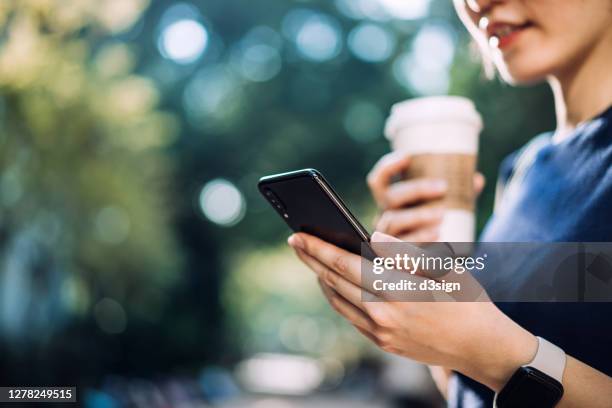 close up of young asian businesswoman taking a coffee break in urban park. she is using smartphone and drinking a cup of coffee, with green nature in the background - paid search stock pictures, royalty-free photos & images